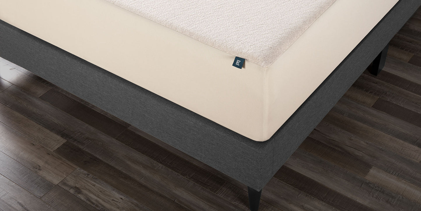Keetsa waterproof mattress protector fitted on mattress with dark bed frame, showing cream hemp top surface with snug ribbed skirt wrapping around corners on dark wood floor.