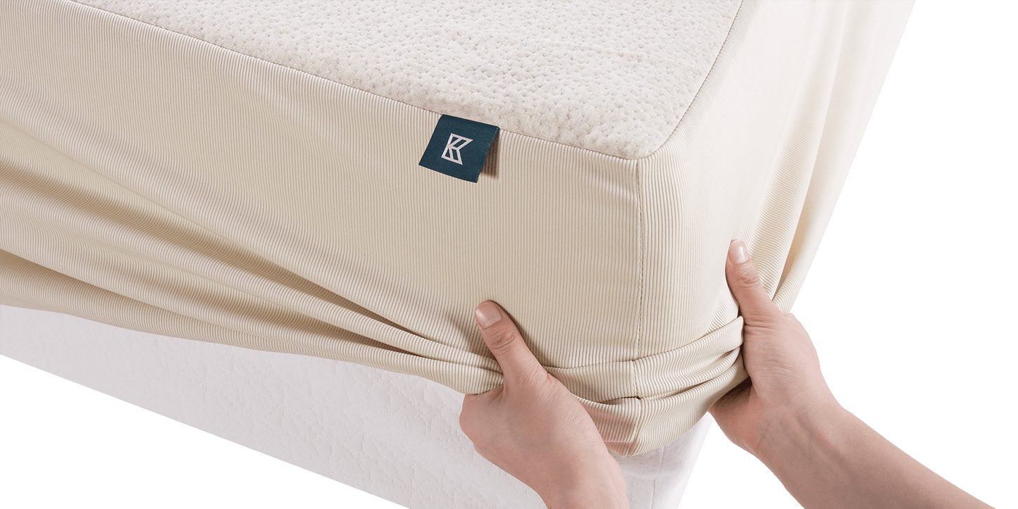 Keetsa waterproof mattress protector being fitted onto mattress by hand, showing stretchy ribbed skirt with deep pocket design, branded Keetsa tag, and textured hemp top surface.