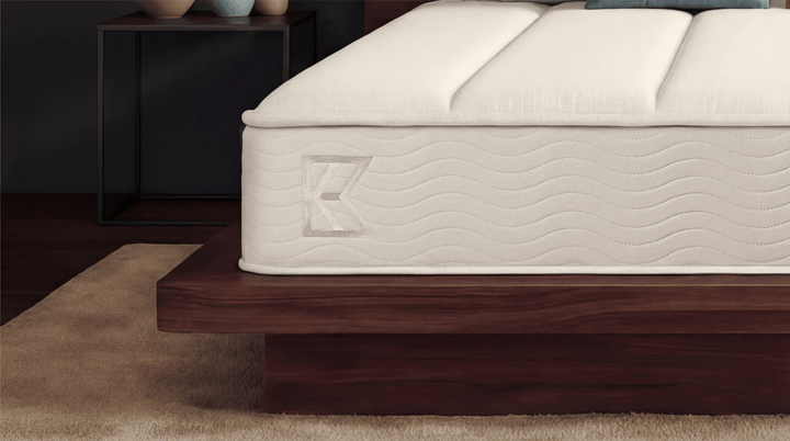 Detailed side view of Keetsa Plus mattress edge with stitched wave pattern and subtle K logo on a solid wood platform, emphasizing edge support, textured fabric, and quality craftsmanship for long-lasting performance.