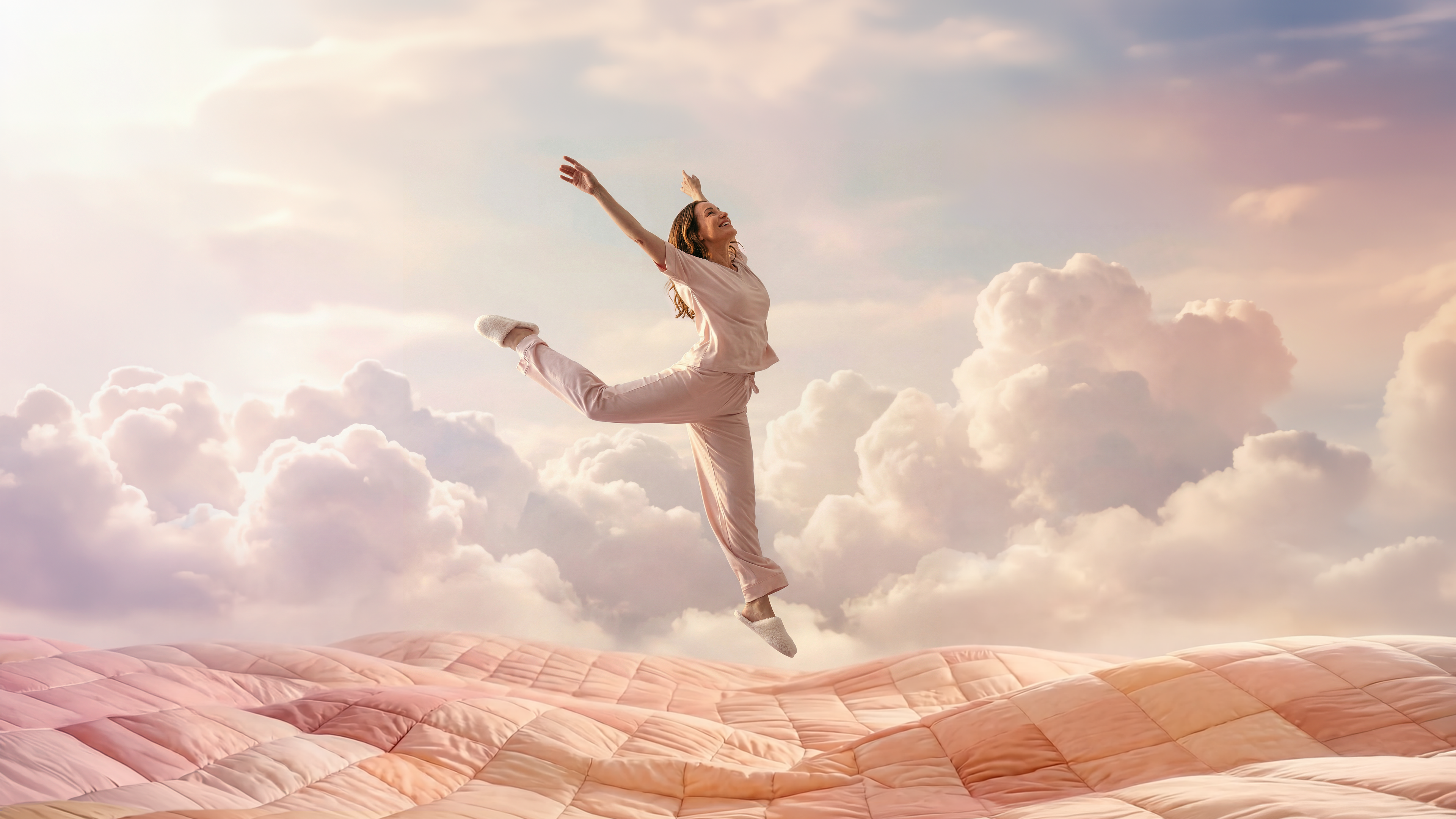 Woman in soft pink pajamas leaps joyfully with arms raised on a vast quilted patchwork comforter shaped like rolling hills, set against a dreamy sky of pastel clouds. Whimsical scene captures the Keetsa feeling of waking up refreshed.