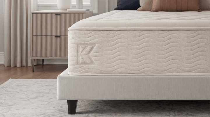 Keetsa Dream mattress foot-of-bed angle showing diamond-quilted sleep surface, wave-stitched side panel with embossed Keetsa logo, and upholstered platform base on marble-patterned rug in neutral bedroom
