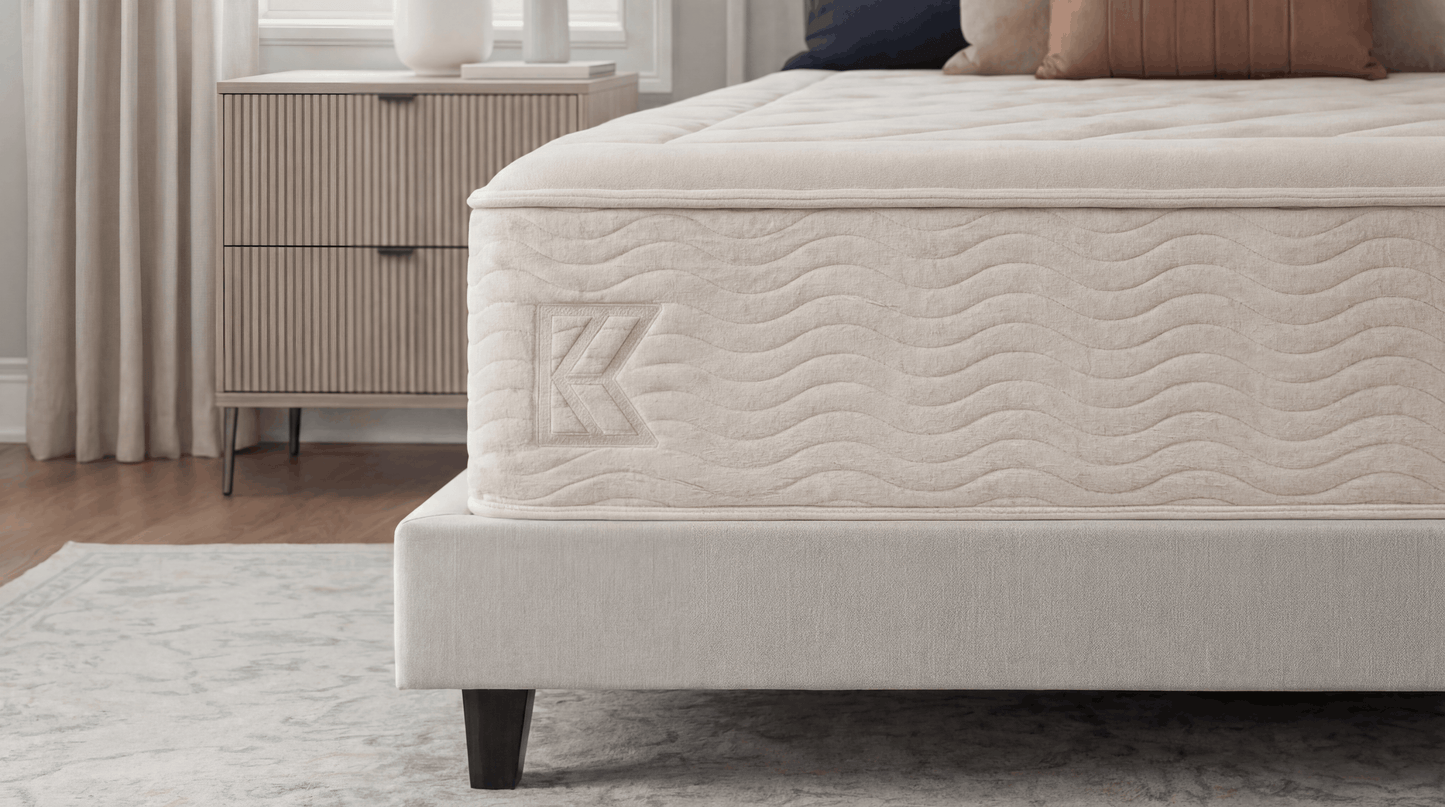 Keetsa Dream mattress foot-of-bed angle showing diamond-quilted sleep surface, wave-stitched side panel with embossed Keetsa logo, and upholstered platform base on marble-patterned rug in neutral bedroom