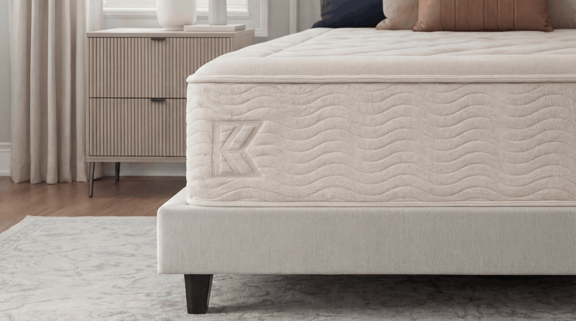 Keetsa Dream mattress foot-of-bed angle showing diamond-quilted sleep surface, wave-stitched side panel with embossed Keetsa logo, and upholstered platform base on marble-patterned rug in neutral bedroom