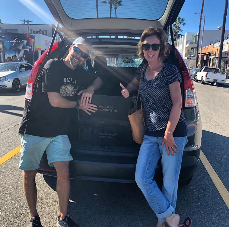 Smiling couple loading a Keetsa mattress box into the back of their SUV outside the store
