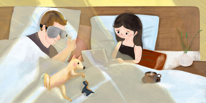 Keetsa eye mask lifestyle illustration of couple in bed with one person sleeping wearing the gray eye mask while partner works on laptop with dog and coffee nearby.
