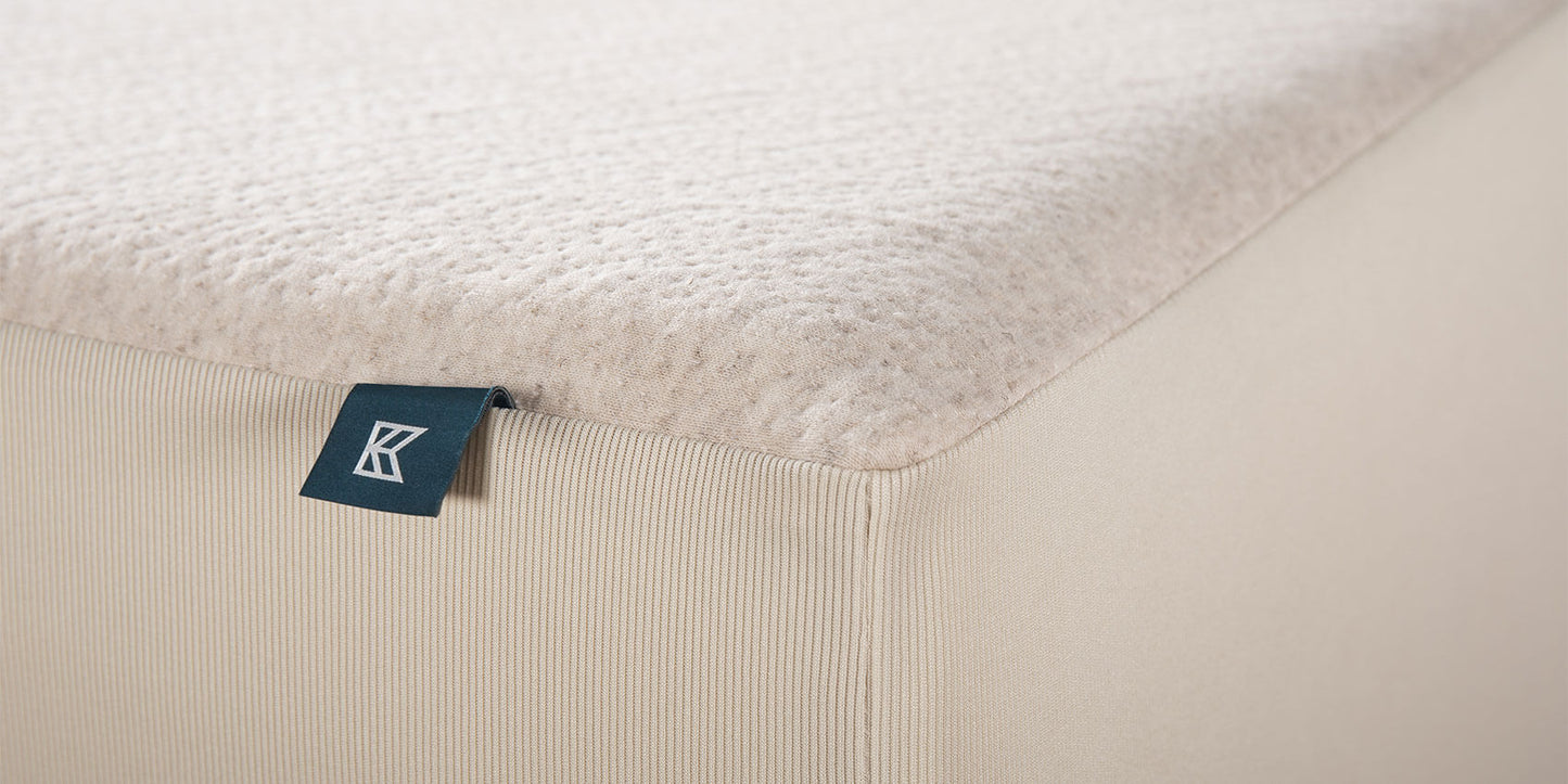 Keetsa waterproof mattress protector close-up showing natural hemp top with textured weave, ribbed fitted skirt in cream, and branded Keetsa tag at the corner seam.
